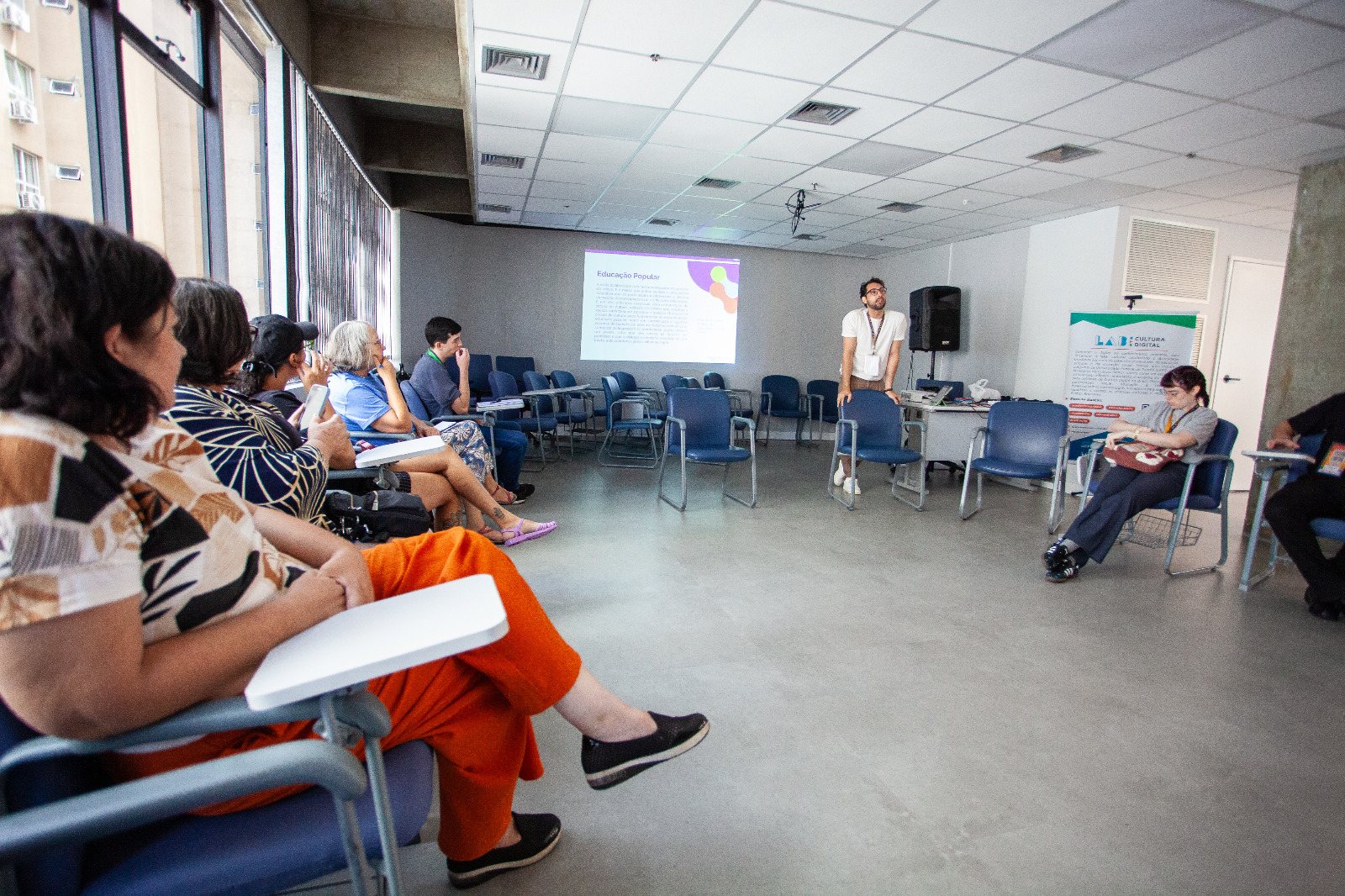 Turma sentada em roda acompanha uma apresentação sobre educação popular em sala com projetor