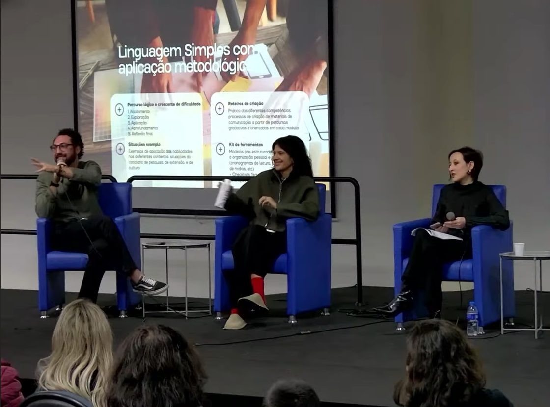 Três integrantes da equipe em poltronas azuis diante de um telão sobre linguagem simples